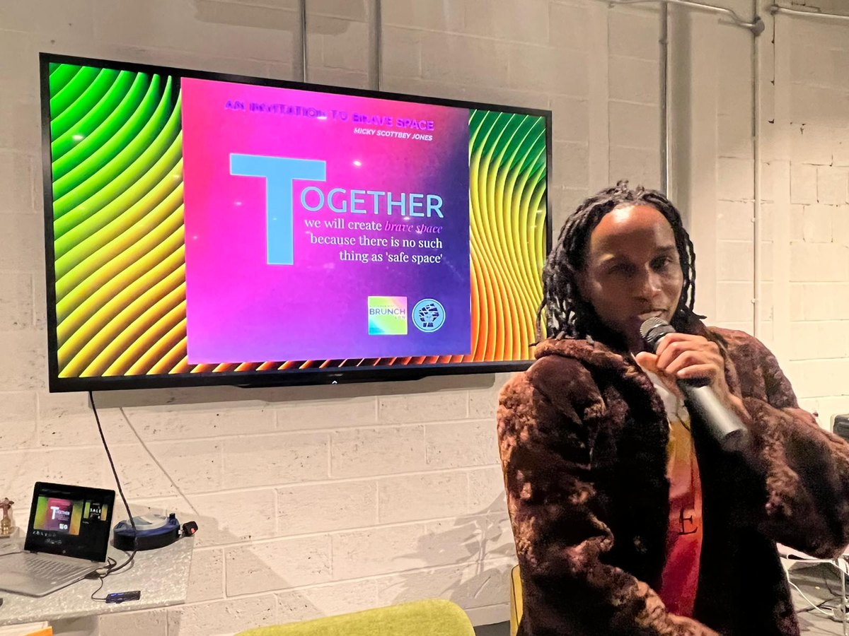 A member speaking at an event beneath a screen reading: together we will create brave space